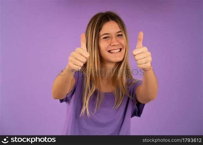 Young woman showing thumbs up to camera and smiling while standing against isolated background.