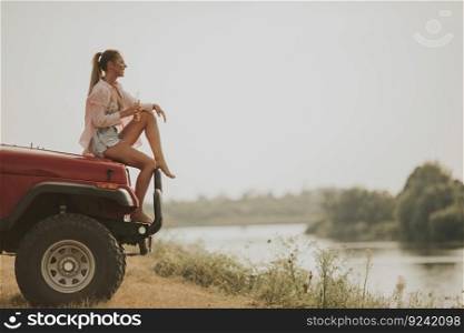 Young woman sits on a car hood,  drinks refreshment and enjoys it