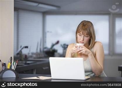 Young woman sits with cup at laptop in kitchen