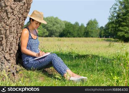 Young woman sitting against tree trunk writing in meadow