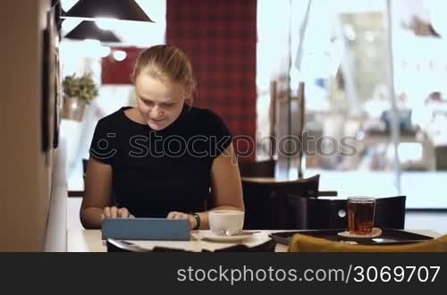Young woman sitting alone in cafe and typing on her tablet while drinking coffee