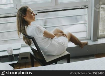 Young woman sitting and daydreaming on a chair by the window in her nightgown