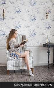 young woman sitting arm chair against wallpaper reading book