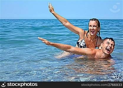 young woman sitting astride man in sea near coast, lifted hands upwards