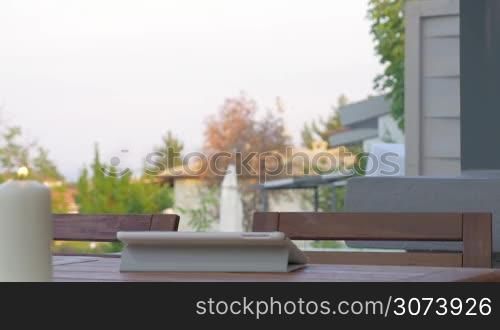 Young woman sitting at the wooden table in the backyard of the rented house. She working with tablet computer and drinking water. Hiring a house, hostel or villa for holidays