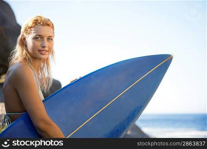 Young woman sitting by rocks holding surfboard
