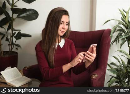 Young woman sitting in a red armchair with cell phone. Opened book and home plants on the background.