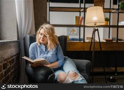 Young woman sitting in chair and reading a book. Female person reads in comfortable couch, living room interior on background