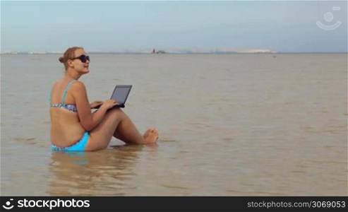 Young woman sitting in the sea with laptop on the knees and typing