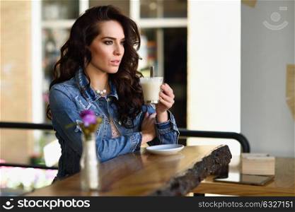 Young woman sitting indoor in trendy urban cafe looking through the window. Cool young modern caucasian female model in her 20s. Cafe city lifestyle.