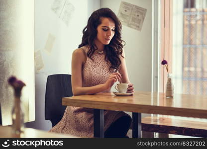 Young woman sitting indoor in trendy urban cafe moving teaspoon into coffee cup. Cool young modern caucasian female model in her 20s.