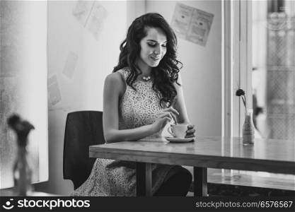 Young woman sitting indoor in trendy urban cafe moving teaspoon into coffee cup. Cool young modern caucasian female model in her 20s.