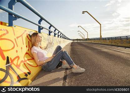 Young woman sitting leaning against graffiti wall on bridge in sunlight