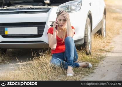 Young woman sitting next to broken car and calling for assistance