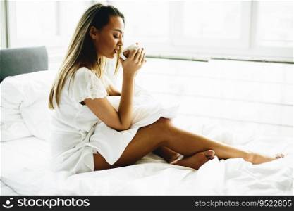 Young woman sitting on a bed and drinking coffee after waking up
