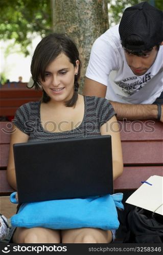 Young woman sitting on a bench and using a laptop with a young man leaning behind her