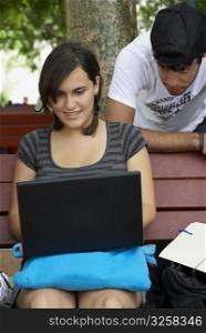 Young woman sitting on a bench and using a laptop with a young man leaning behind her