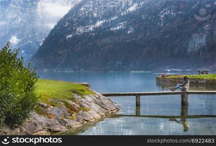 Young woman sitting on a wooden deck, relaxing and admiring the majestic Dachstein Mountains and the Hallstatter lake, located in Hallstatt, Austria.