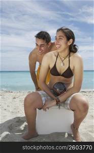 Young woman sitting on an ice box on the beach with a young man standing behind her