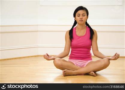 Young woman sitting on floor meditating in yoga lotus pose with legs crossed and eyes closed.