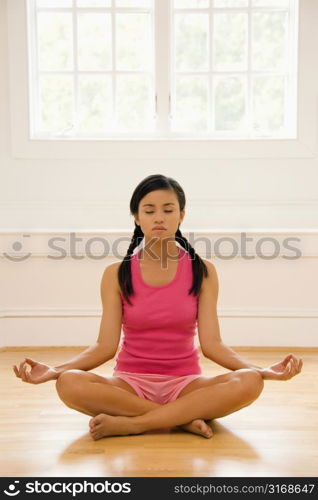 Young woman sitting on floor meditating in yoga lotus pose with legs crossed and eyes closed.