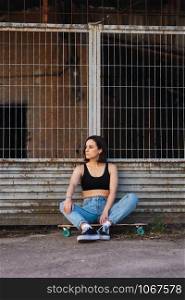 Young woman sitting on her skate in an old industrial street leaning against metal fence