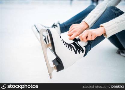 Young woman sitting on ice and ties the shoelaces on skates, skating rink. Winter ice-skating on open air, active leisure. Young woman ties the shoelaces on skates