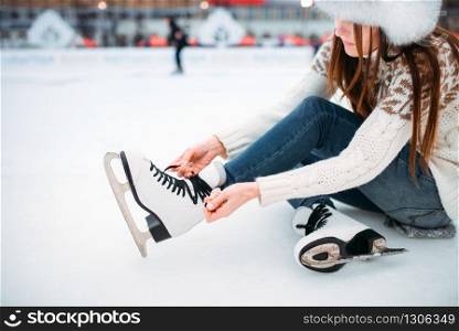 Young woman sitting on ice and ties the shoelaces on skates, skating rink. Winter ice-skating on open air, active leisure