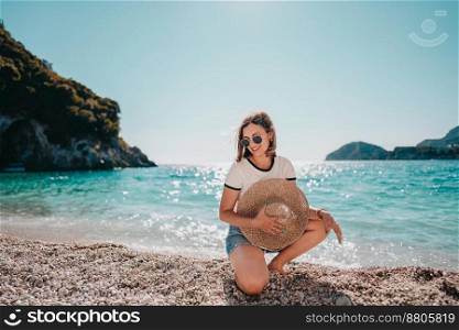Young woman sitting on pebble beach near Mediterranean Ionian sea. Lady tourist enjoying vacation time, holding straw hat and smiling, nature background. . High quality photo. Young woman sitting on pebble beach near Mediterranean Ionian sea. Lady tourist enjoying vacation time, holding straw hat and smiling, nature background. 