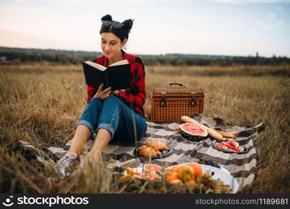 Young woman sitting on plaid and reads book, picnic in summer field. Romantic junket, happy holiday