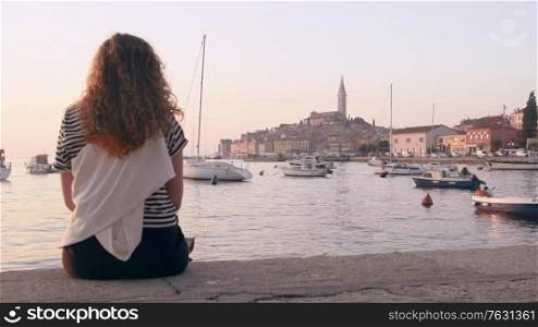 Young woman sitting on Rovinj marina and enjoying city view with boats and St. Euphemia cathedral at sunset. Istria, Croatia.. Young woman taking a picture of Rovinj