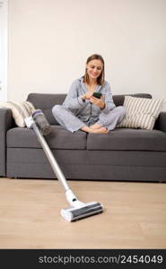 Young woman sitting on sofa and using mobile phone while cleaning at home, copy space. Housework and chores concept. Cleaning appliances.. Young woman sitting on sofa and using mobile phone while cleaning at home, copy space. Housework and chores concept. Cleaning appliances