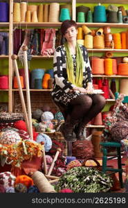 Young Woman Sitting On Stool Holding Knitting Needles In Front Of Yarn Display