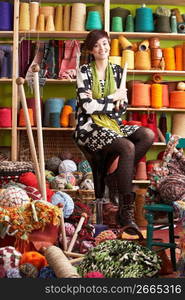 Young Woman Sitting On Stool Holding Knitting Needles In Front Of Yarn Display