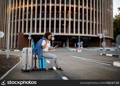 Young woman sitting on suitcase at the entrance to car parking. Female traveler with luggage near vehicle park lot, passenger with bag. Girl with baggage on city street. Woman sitting on suitcase, entrance to car parking