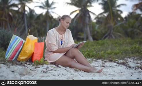 Young woman sitting on the beach and using touch pad, shopping bags nearby. Vacation with pad