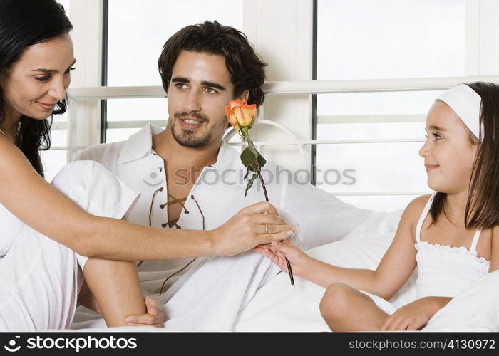 Young woman sitting on the bed giving a flower to her to her daughter with a young man sitting beside her