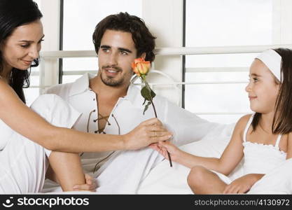 Young woman sitting on the bed giving a flower to her to her daughter with a young man sitting beside her