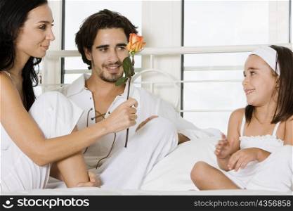 Young woman sitting on the bed holding a flower with a young man and her daughter