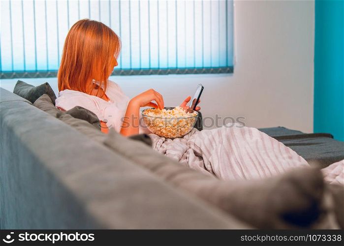 Young woman sitting on the bed sofa at home, lying with a popcorn bowl covered with blanket at her apartment alone enjoying using mobile smart phone to take selfies photo or to send sms message