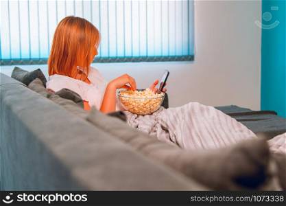 Young woman sitting on the bed sofa at home, lying with a popcorn bowl covered with blanket at her apartment alone enjoying using mobile smart phone to take selfies photo or to send sms message