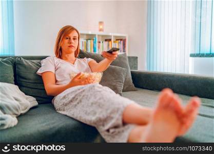 Young woman sitting on the bed sofa at home, lying with a popcorn bowl watching TV covered with blanket at her apartment alone enjoying movies or series holding remote control pointed