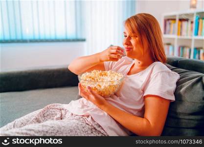 Young woman sitting on the bed sofa at home, lying with a popcorn bowl watching TV covered with blanket at her apartment alone enjoying movies or series
