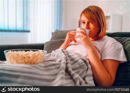 Young woman sitting on the bed sofa at home, lying with a popcorn bowl watching TV covered with blanket at her apartment alone enjoying movies or series having a cup of tea or coffee