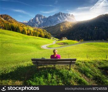 Young woman sitting on the bench and beautiful alpine village at sunset in autumn.Tyrol, Dolomites, Italy. Colorful landscape with girl, green meadows, orange trees, road, mountain, blue sky in fall. Young woman sitting on the bench and beautiful alpine village