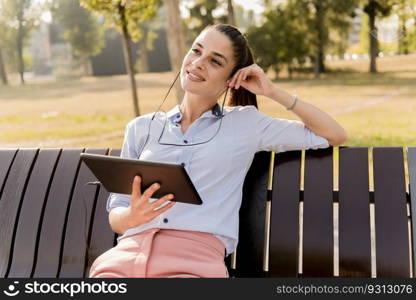 Young woman sitting on the bench in the park and listen music on  a tablet