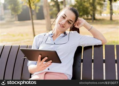 Young woman sitting on the bench in the park and listen music on  a tablet