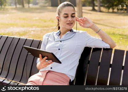 Young woman sitting on the bench in the park and listen music on  a tablet