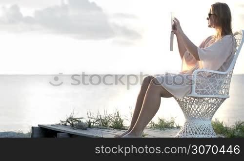 Young woman sitting on the chair by the sea and taking pictures with tablet computer