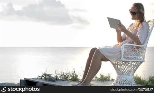 Young woman sitting on the chair by the sea and watching pictures with tablet computer